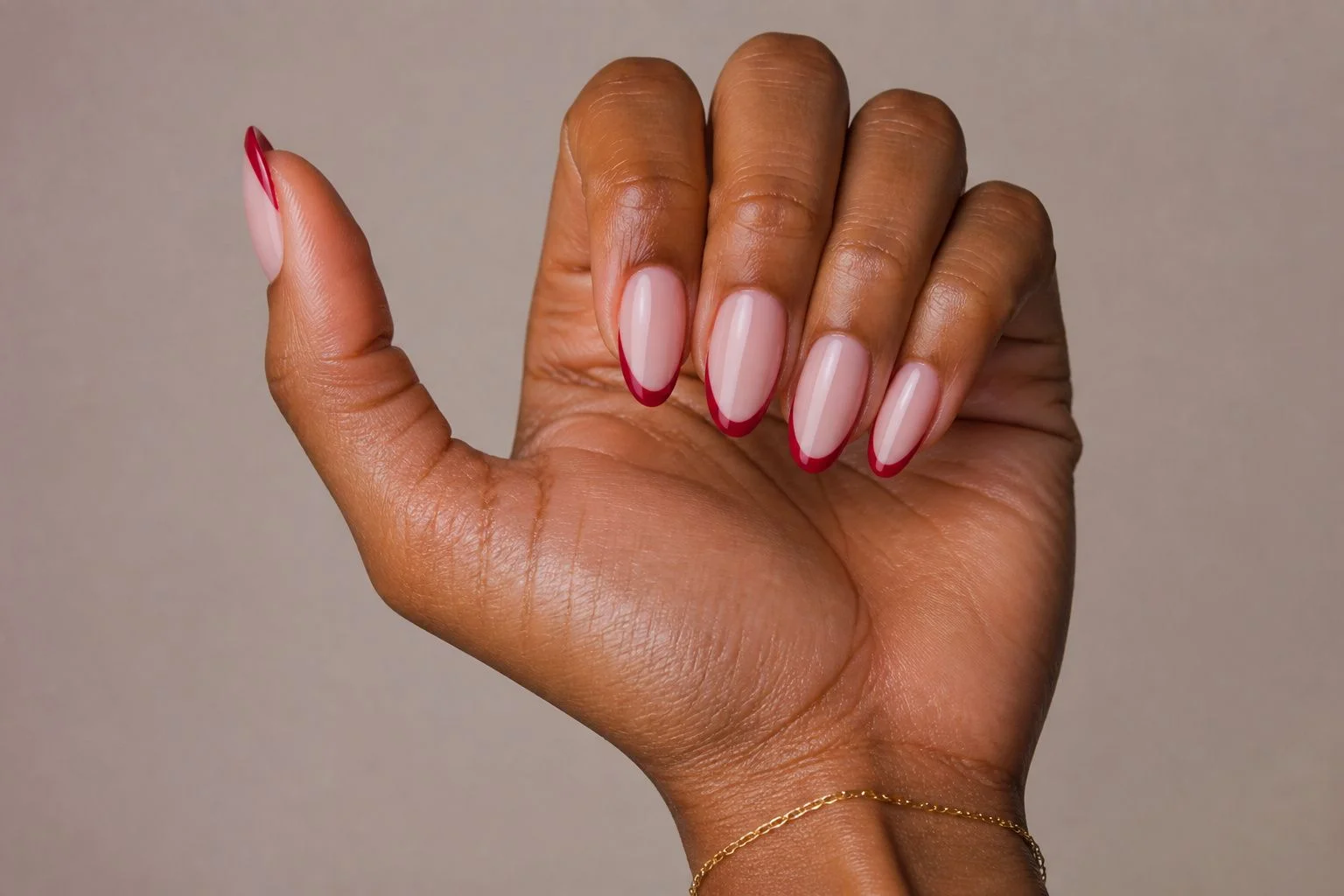 Curled hand showing a glossy nude almond manicure with a classic red French tip accent and a gold bracelet