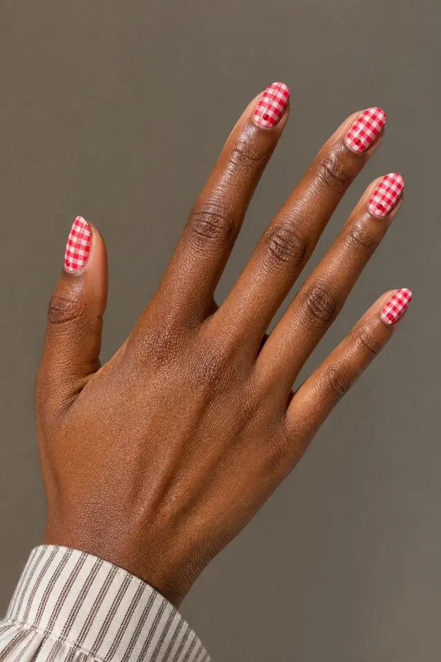 Red gingham with tiny cherry-red dot accents on short squoval nails, glossy finish, deep brown skin, hand turned toward daylight with connected forearm