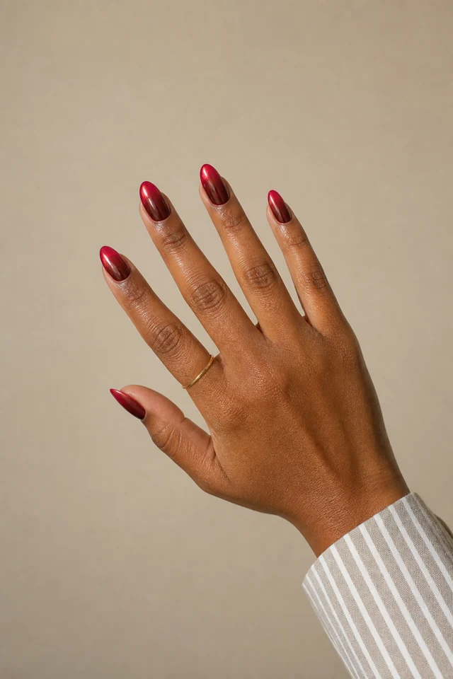 Cherry-red chrome ombre almond nails fading from deep burgundy at the cuticle on a medium-deep brown hand with visible wrist in natural daylight