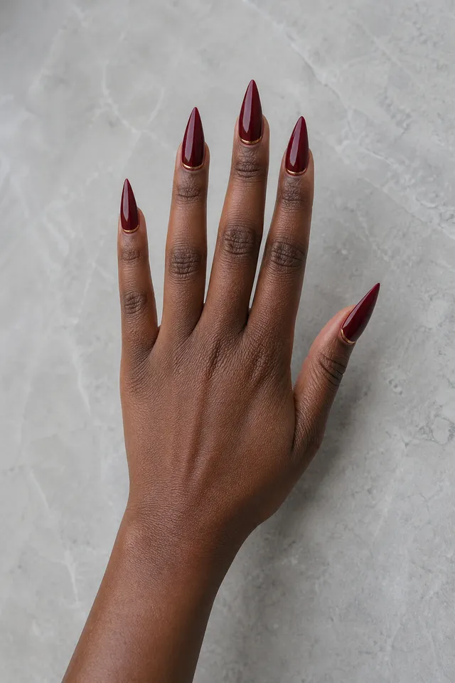 deep burgundy glossy stiletto nails on a deep brown hand posed palm-down on a pale surface with visible forearm
