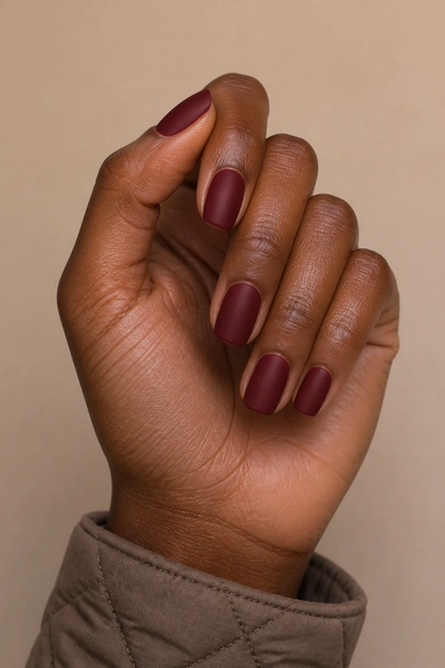 Burgundy red matte nails on a rich dark brown skin tone hand in a lightly crossed fingertip pose with visible wrist and short partial forearm, natural daylight
