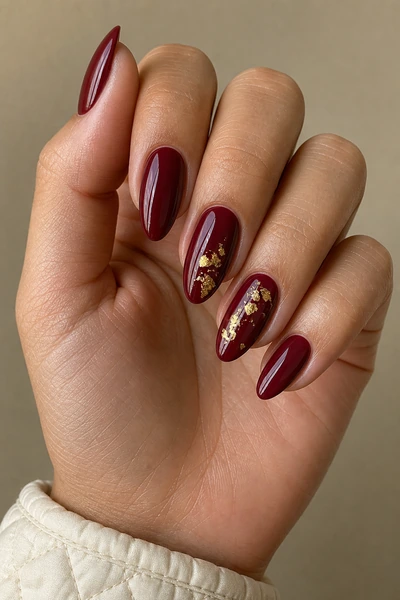 Burgundy nails with gold foil accents on a medium olive skin tone hand in a softly curled pose with visible wrist and short partial forearm, natural daylight
