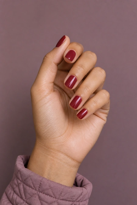 Short glossy cranberry red nails on a light brown skin tone hand in a gentle pinch pose with visible wrist and short partial forearm, natural daylight