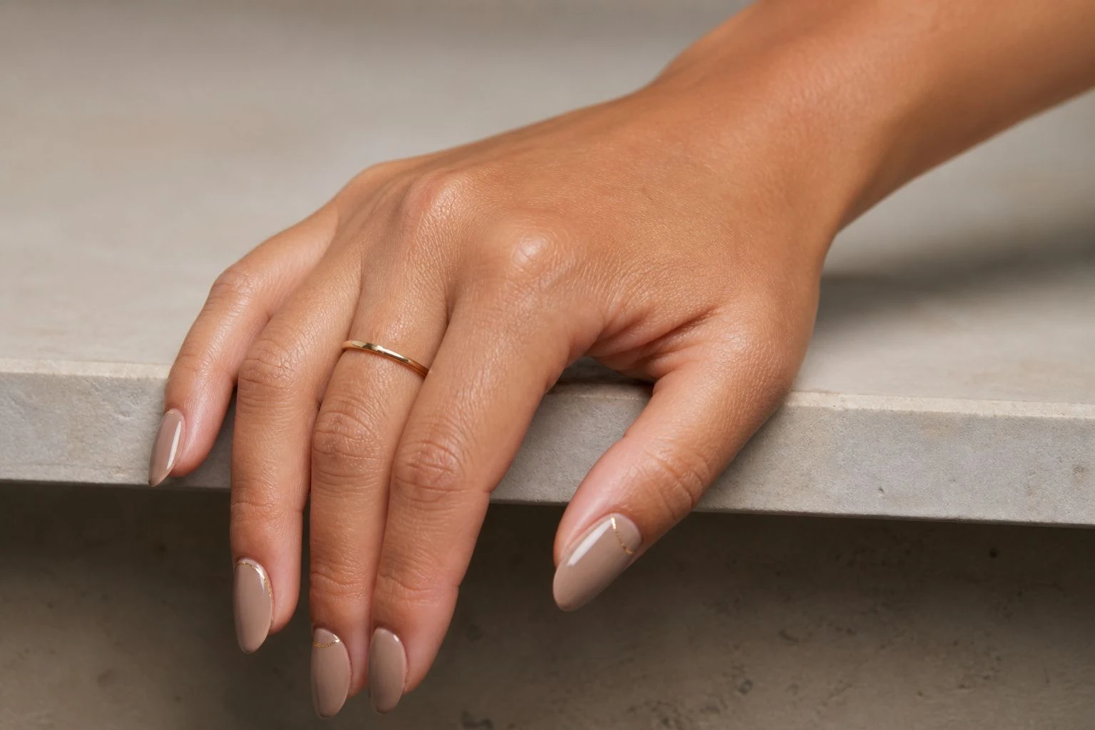 Warm taupe nude nails with a tiny gold detail and a delicate gold ring, table edge pose