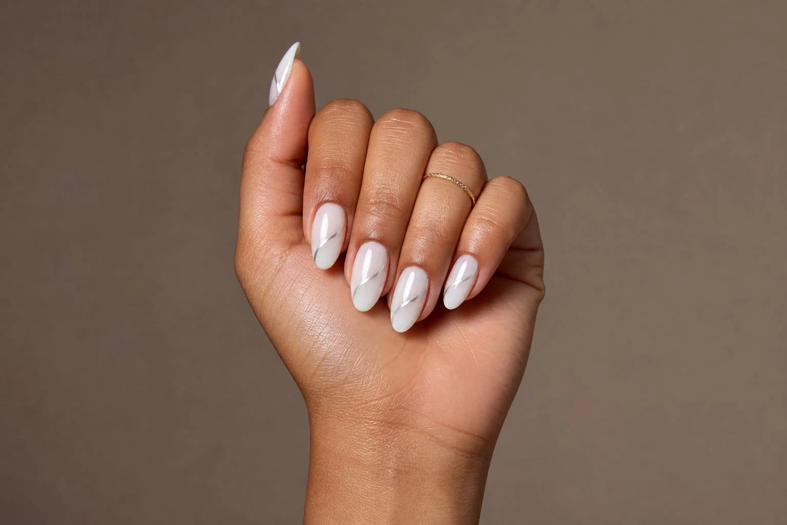Curled hand showing a glossy white almond manicure with a thin silver line accent and a delicate gold ring