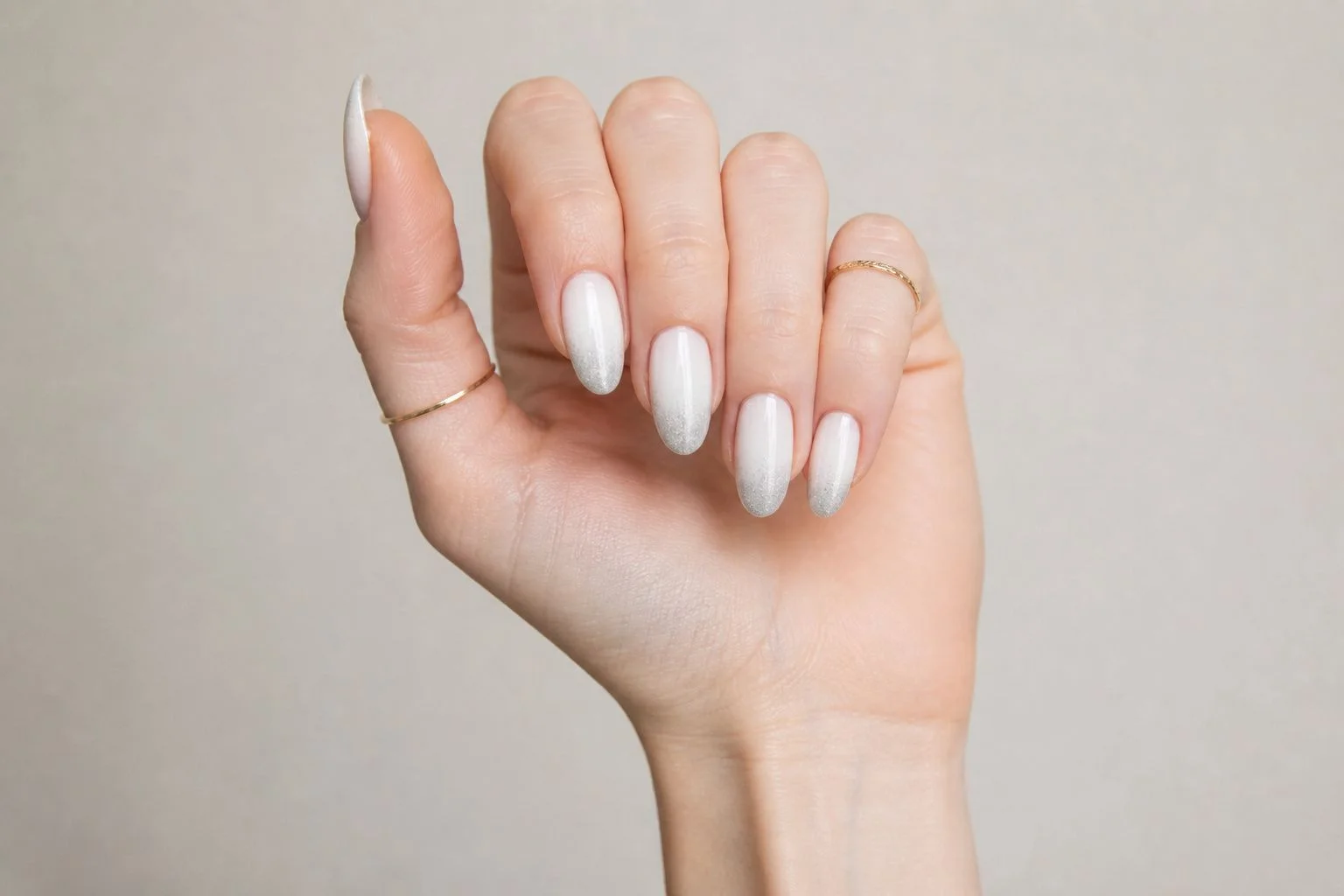 Curled hand showing a soft white almond manicure with a delicate silver ombre fade and two thin gold rings