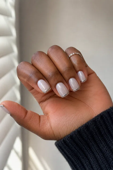 Milky white chrome short squoval nails with delicate silver French lines on a deep brown skin tone, shown on one side-angled hand with a dark cuff and connected short wrist