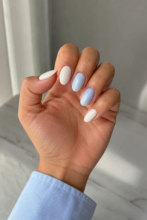 Creamy white short almond nails with icy blue shimmer accents on a medium tan skin tone, shown on one palm-up fingertip-rest hand with a pale blue cuff and connected short wrist