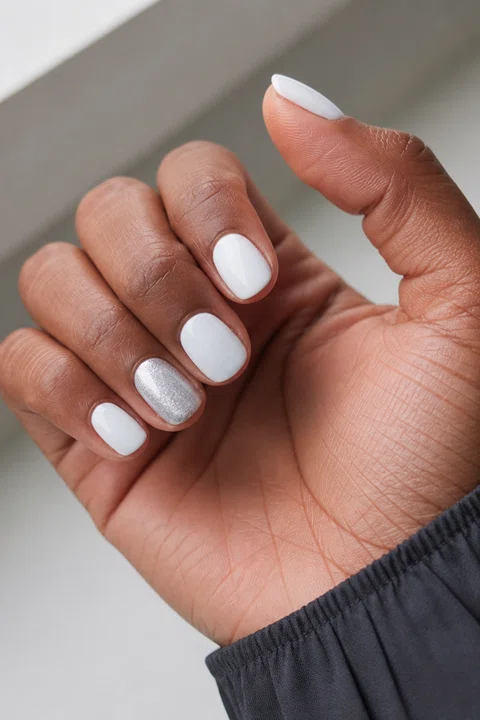 Glossy crisp white short oval nails with a silver shimmer accent on a rich dark brown skin tone, shown on one palm-up hand with a charcoal cuff and connected short wrist