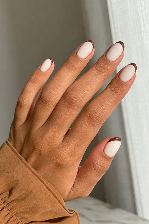 Creamy white short almond nails with cocoa-brown French edges and a gold shimmer accent on a medium warm brown skin tone, shown on one tilted hand with a camel cuff and connected short wrist