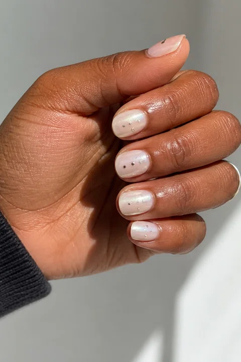 Sheer white glossy short oval nails with pearl shimmer and tiny silver dot accents on a rich dark brown skin tone, shown on one low curled hand with a charcoal cuff and connected short wrist