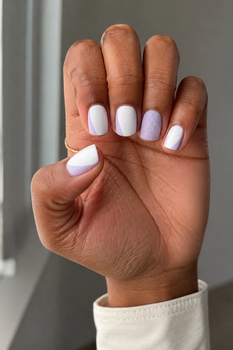 Snowy white short squoval nails with icy lavender side sweeps and one matte accent on a medium-deep brown skin tone, shown on one upright hand with a cream shirt cuff and connected short wrist