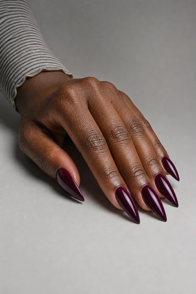 Long plum acrylic stiletto nails on a fingertip-rest hand pose with visible forearm, deep brown skin tone, natural daylight