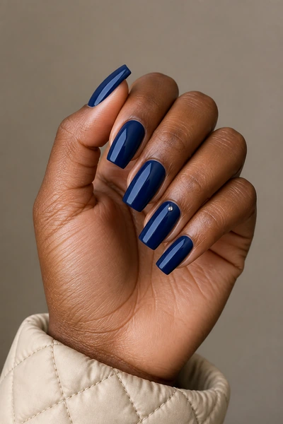Sapphire acrylic nails with minimal jewel accents on a softly curled hand pose with visible wrist, deep brown skin tone, soft daylight