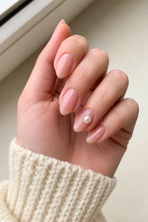 Glossy sheer blush nude medium-short almond nails with one pearl accent on fair skin, shown on one relaxed three-quarter hand with a cream knit cuff and connected short wrist