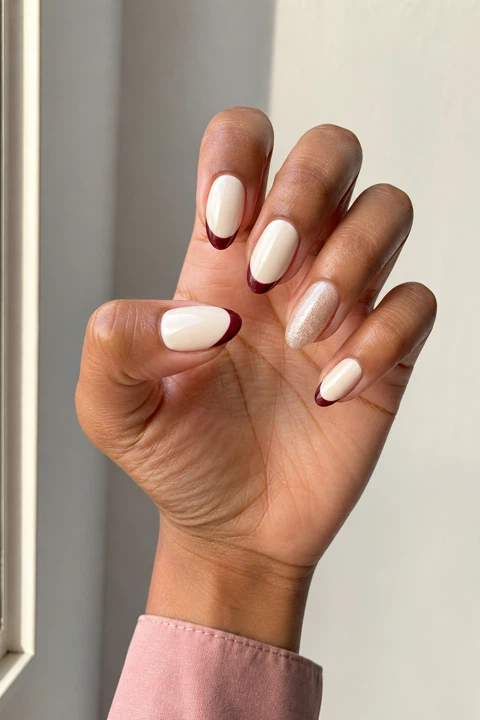 Glossy creamy ivory almond nails with berry French tips and one shimmer accent on medium warm brown skin, shown on one upright fan hand with a blush shirt cuff and connected short wrist