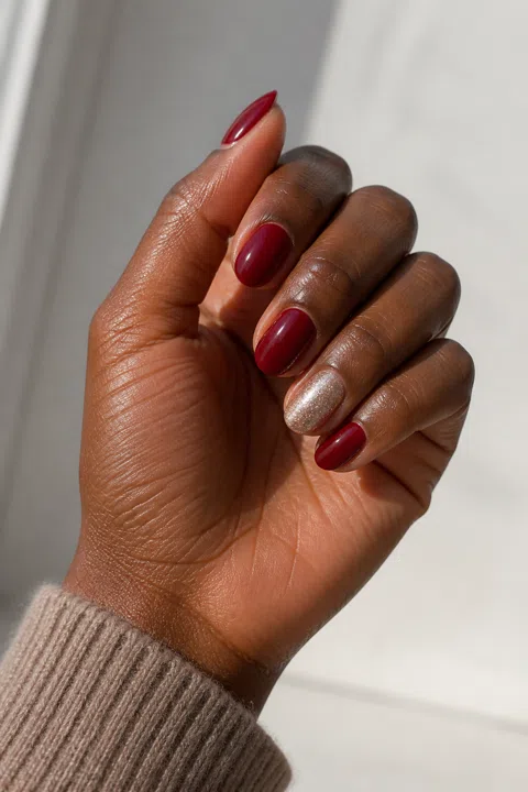 Glossy cranberry red short almond nails with one rose-gold shimmer accent on deep ebony brown skin, shown on one gently curled palm-up hand with a taupe knit cuff and connected short wrist
