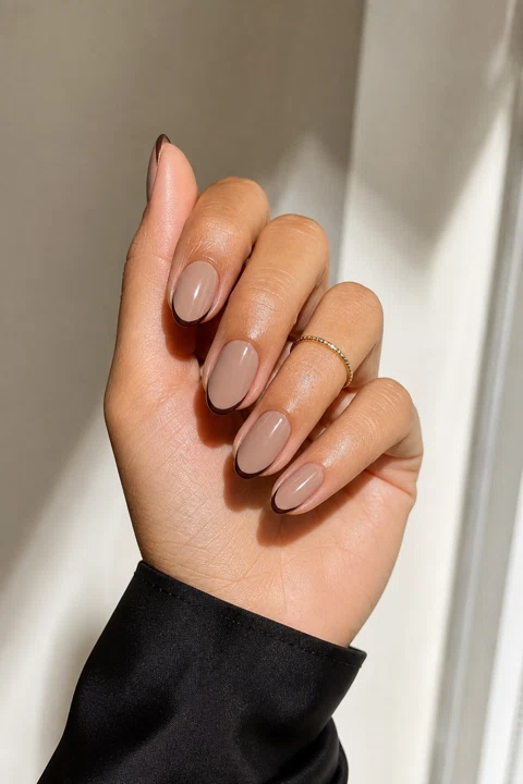 Glossy warm taupe nude short almond nails with deep espresso micro-French tips on medium tan olive skin, shown on one three-quarter hand with a black satin sleeve, thin gold ring, and connected short wrist