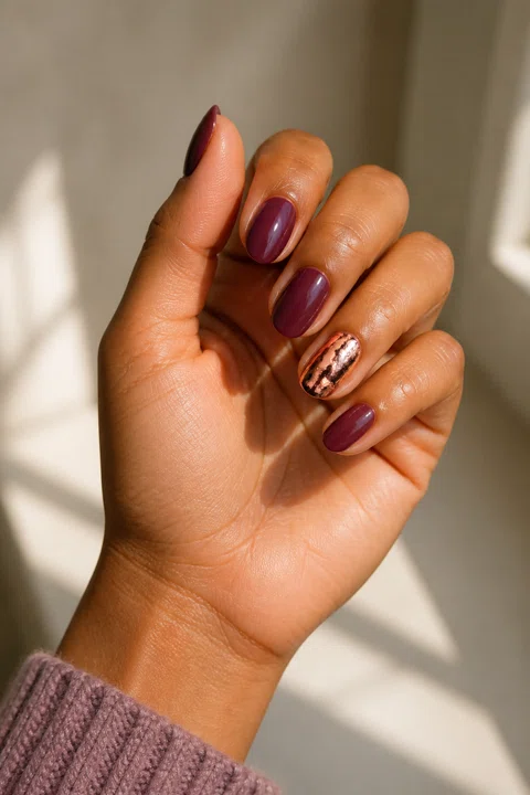 Glossy rich plum short almond nails with one rose-gold foil accent on medium warm caramel skin, shown on one gently curled palm-up hand with a mauve knit cuff and connected short wrist