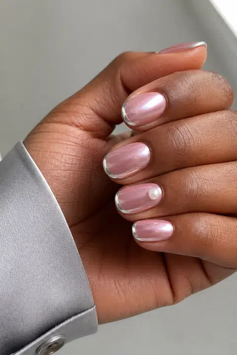 Icy rose chrome short oval nails with slim silver French edges and a pearl accent on a rich dark brown skin tone, shown on one low curled hand with a grey cuff and connected short wrist