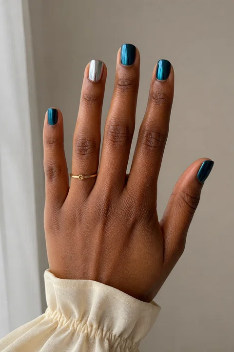 Deep teal chrome short squoval nails with one silver mirrored accent on a medium-deep brown skin tone, shown on one upright hand with a cream cuff and connected short wrist