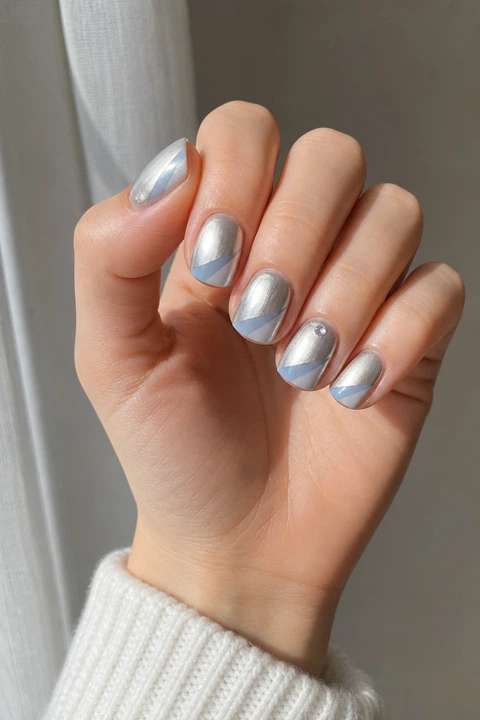 Frosted silver chrome short rounded-square nails with pale blue diagonal accents on a fair skin tone, shown on one relaxed hand with a white knit cuff and connected short wrist