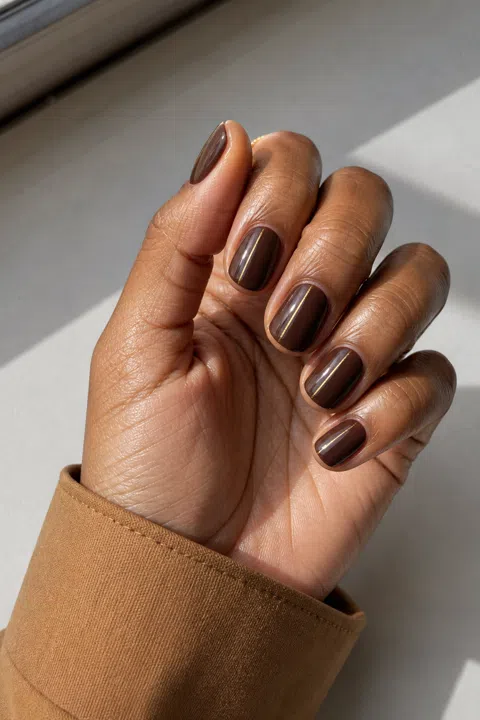 Glossy warm espresso gel short oval nails with a tiny gold line accent on a deep brown skin tone, shown on one palm-up fingertip-rest hand with a camel cuff and connected short wrist
