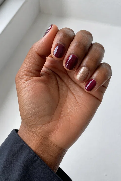 Glossy warm burgundy gel short oval nails with one champagne shimmer accent on a deep brown skin tone, shown on one softly curled palm-up hand with a charcoal cuff and connected short wrist
