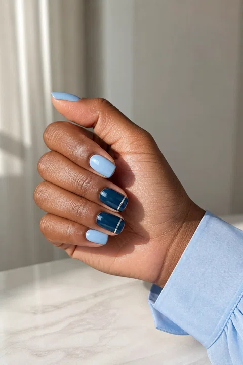 Glossy icy blue gel short squoval nails with deep navy accents and a fine silver line on a medium-deep brown skin tone, shown on one side-angled hand with a pale blue cuff and connected short wrist