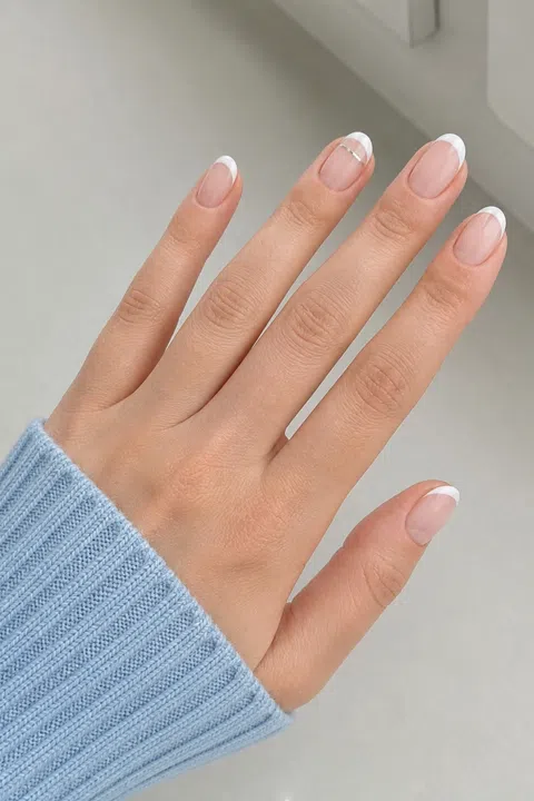 Sheer nude gel short almond nails with crisp white French tips and one silver line accent on a fair skin tone, shown on one palm-down relaxed hand with a pale blue cuff and connected short wrist