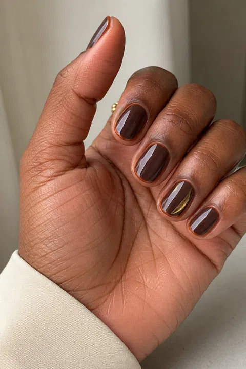 Glossy chocolate gel short oval nails with caramel micro-French tips and one gold accent on a rich dark brown skin tone, shown on one palm-up fingertip-rest hand with a cream cuff and connected short wrist