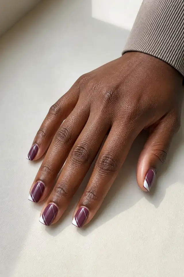 Warm berry-plum glossy medium square nails with creamy ivory diagonal tips and silver pinstripes on deep rich brown skin, shown in a palm-down side-angled fingertip-rest pose with a short partial wrist