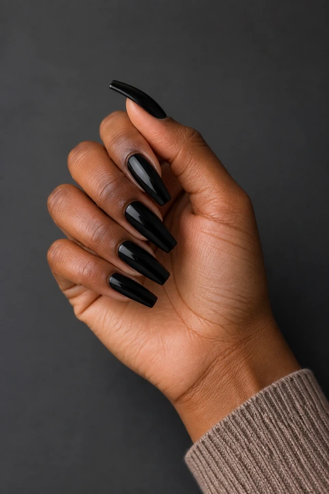 Glossy black moderate-length coffin nails on deep brown skin in an upright softly curled pose with a taupe knit sleeve, visible short wrist, and natural daylight