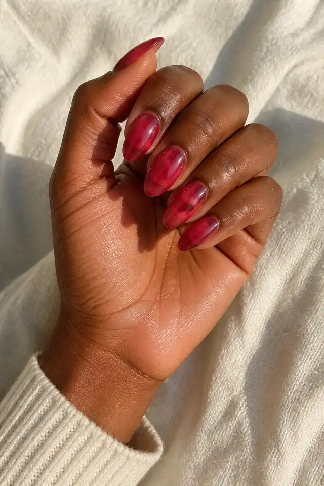 Soft berry red acrylic almond nails with rich dark brown skin, a gently curled diagonal hand, and a cream sleeve in natural daylight