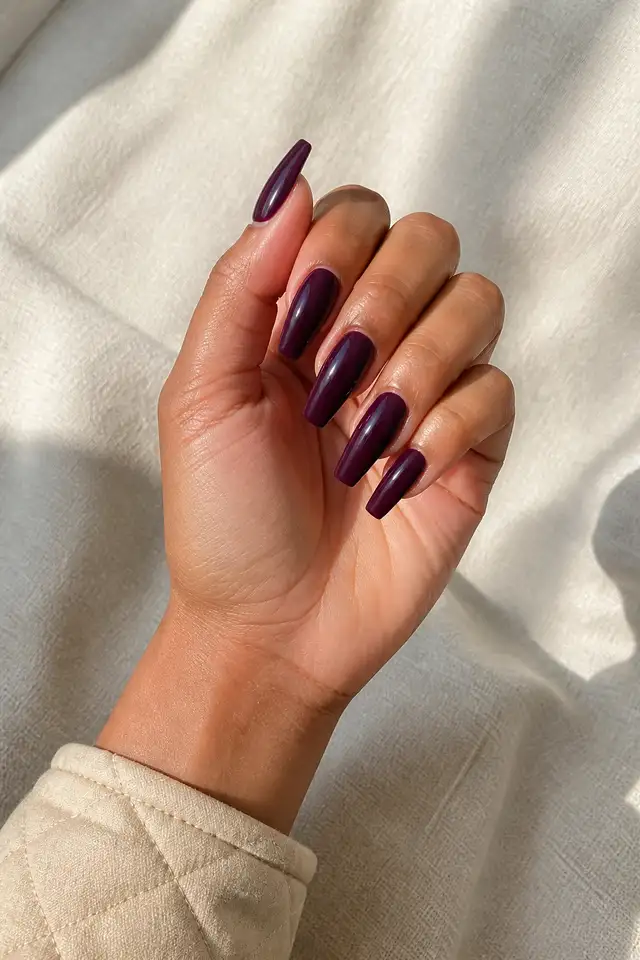 Deep plum glossy acrylic coffin nails with medium brown skin, a softly curled hand, and a beige quilted cuff in natural daylight
