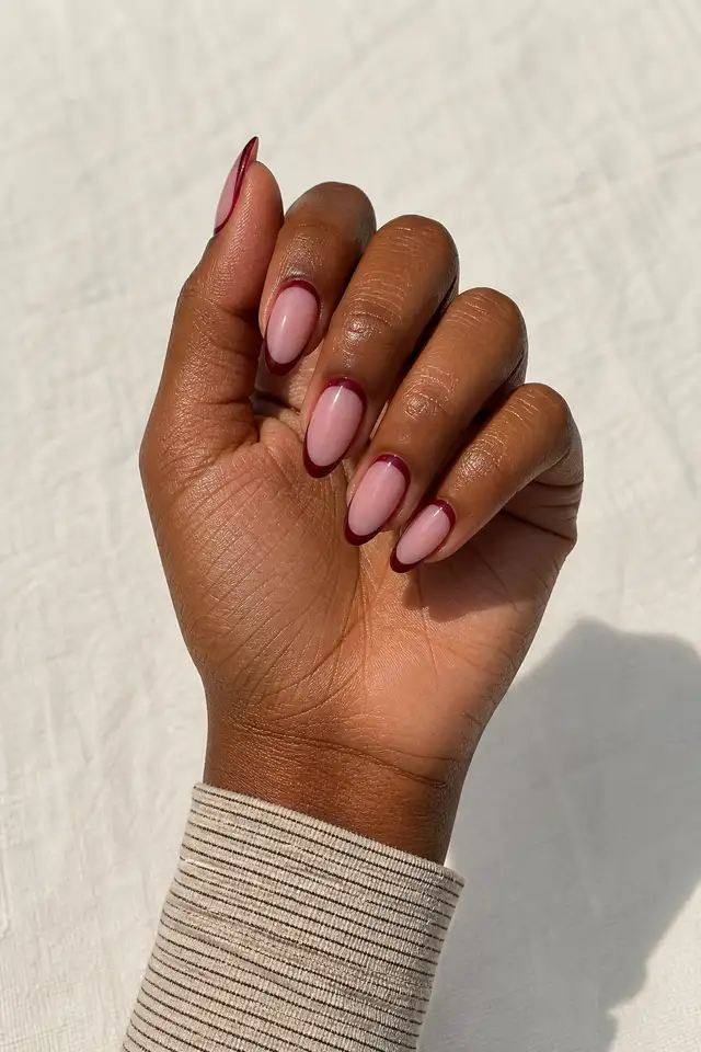 Sheer pink and burgundy reverse French acrylic almond nails with deep brown skin, a compact curled pose, and a narrow sleeve in natural daylight