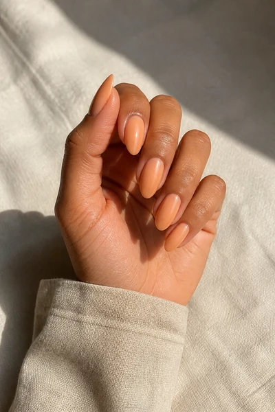 Peach beige almond nails with a glossy glazed finish on a tan skin tone hand in a relaxed curled pose with a beige linen cuff and visible wrist