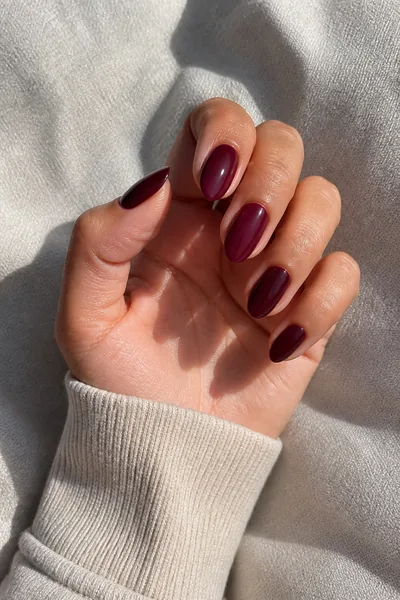 Deep wine almond nails with a glossy finish on a tan skin tone hand in a gentle curled pinch pose with an oatmeal cuff and visible wrist