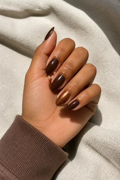 Glossy espresso almond nails with one subtle bronze accent on a light brown skin tone hand in a compact curled pose with a mocha sleeve and visible wrist