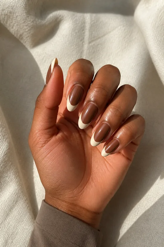 Deep brown skin hand in a compact curled pose wearing glossy cocoa almond nails with creamy ivory French tips and a muted sleeve cuff