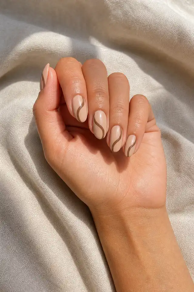 Latte beige glossy medium oval nails with curved espresso brown swirls and one creamy nude accent on medium neutral tan skin, shown in a gentle bent-finger pose with a short partial wrist and no sleeve
