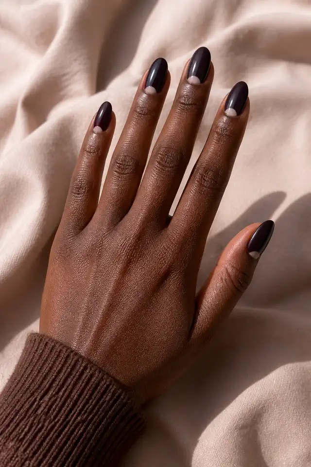 Blackened plum brown glossy medium almond nails with nude half moons and one deep burgundy accent on dark ebony brown skin, shown in a three quarter back of hand pose with a cocoa knit cuff and short partial forearm