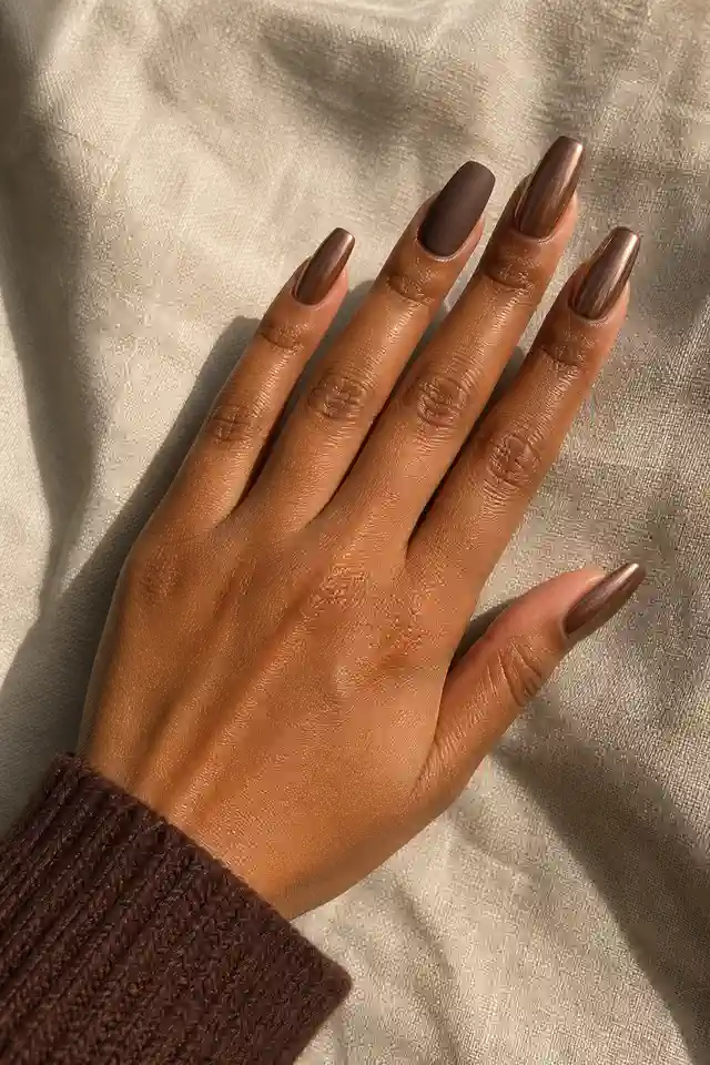 Brown sugar bronze chrome medium coffin nails with one matte cocoa accent on medium deep golden brown skin, shown in a low diagonal back of hand sweep with a cocoa knit cuff and short partial forearm