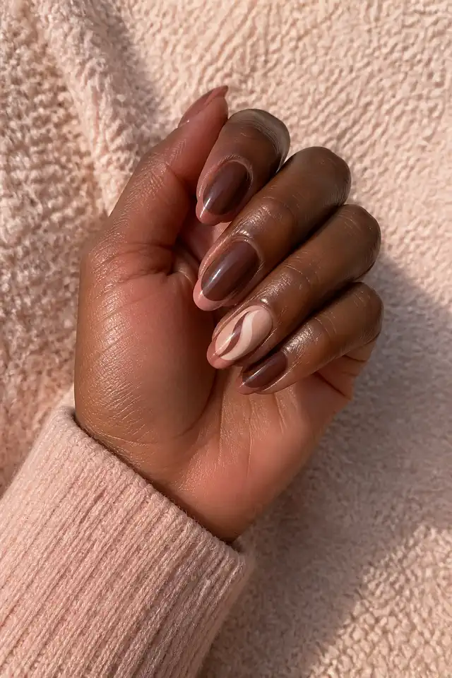 Rosewood brown glossy medium oval nails with thin dusty rose French tips and one cream negative space accent on deep cool brown skin, shown in a compact curved pose with a rose beige sweater cuff and short partial forearm