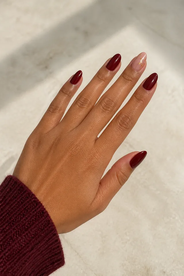 Deep garnet glossy medium almond nails with one sheer blush accent and tiny gold foil flecks on medium tan golden skin, shown in a palm down diagonal pose with a burgundy knit cuff