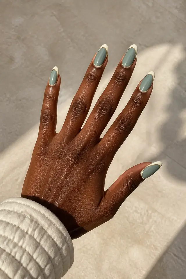Soft sage gray glossy medium almond nails with creamy ivory French tips and a fine gold line on deep rich brown skin, shown in a palm down open fan pose with an ivory quilted cuff