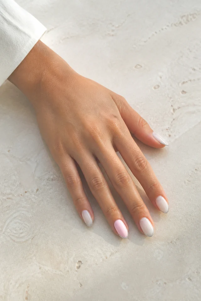 Warm cocoa glossy medium almond nails with caramel nude negative space arcs and one rose gold accent on medium deep neutral brown skin, shown in a side angled fingertip rest pose with a taupe cuff
