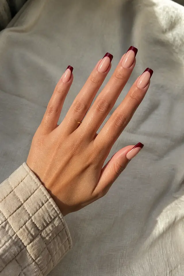 Light olive skin hand with burgundy French tips on medium coffin nails, a diagonal display pose, a beige quilted cuff, and one tiny plain ring in daylight