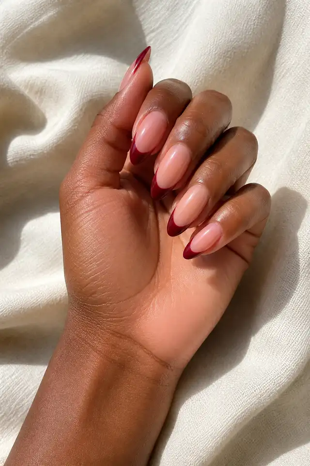 Deep brown skin hand with wine red French tips on long almond nails, a layered fan pose, and a narrow burgundy sleeve in daylight