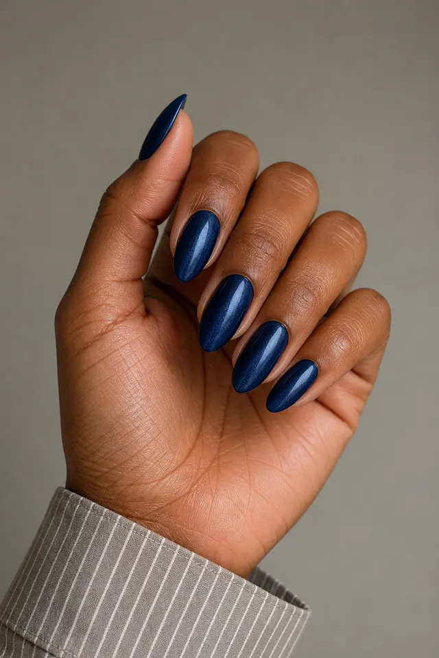 Navy ink velvet almond nails on a gently curled hand with rich dark brown skin and a narrow cream sleeve in natural daylight